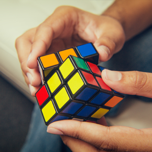 Hands twisting a scrambled Rubik's Cube with bright yellow, red, blue, and green stickers on a dark surface edge.
